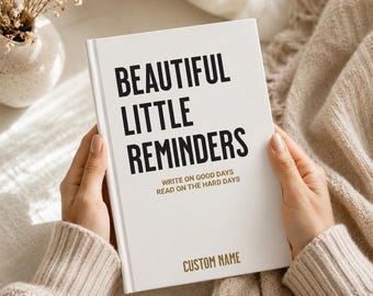 May include: A white hardcover book with the black text BEAUTIFUL LITTLE REMINDERS, the gold text WRITE ON GOOD DAYS READ ON THE HARD DAYS, and the gold text CUSTOM NAME printed on the front cover, held in a pair of hands against a soft fabric.
