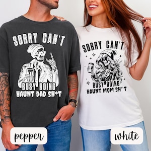 May include: Two t-shirts, one pepper-colored and one white, featuring a skeleton graphic. The text on the shirts reads "SORRY CAN'T BUSY DOING HAUNT DAD SH*T" and "SORRY CAN'T BUSY DOING HAUNT MOM SH*T". The shirts are short-sleeved.