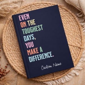 May include: A navy blue journal with the text "EVEN ON THE TOUGHEST DAYS, YOU MAKE A DIFFERENCE." in colorful letters. The words "Custom Name" are written below the main text. The journal is on a woven placemat.