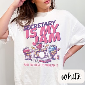 May include: White t-shirt with a pink and purple graphic of fruit playing instruments. The text "Secretary is my jam and I'm here to spread it" is printed on the shirt.