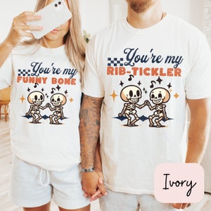 May include: Two white t-shirts with a skeleton graphic and text that reads "You're my funny bone" and "You're my rib-tickler". The shirts are on a couple holding hands. The text is in a vintage style font. The background is a light brown color.