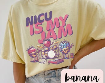 May include: A yellow t-shirt with the text "NICU is my JAM" in pink and black letters. The shirt features a cartoon illustration of fruit playing instruments.