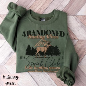 May include: A green sweatshirt with the text "Abandoned Hunting Wives Social Club" and a graphic of a deer in a forest. The text below the graphic says "Hello hunting season goodbye husband."