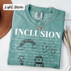 May include: Light green t-shirt with the word "INCLUSION" in large white letters. The shirt features a rainbow, hearts, and other graphics, along with a handwritten message about inclusivity. A silver necklace with a pendant is draped across the shirt.