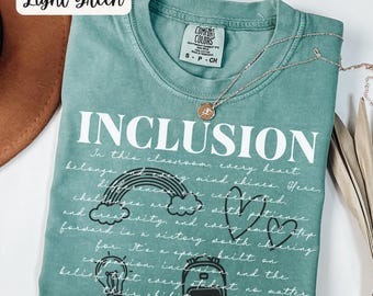 May include: Light green t-shirt with the word "INCLUSION" in large white letters. The shirt features a rainbow, hearts, and other graphics, along with a handwritten message about inclusivity. A silver necklace with a pendant is draped across the shirt.