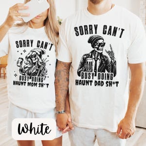 May include: White t-shirts with black graphic designs. One shirt features a skeleton with a beanie, holding a coffee cup, and the text "SORRY CAN'T BUSY DOING HAUNT MOM SH*T." The other shirt has a skeleton in a jacket with a beer and the text "SORRY CAN'T BUSY DOING HAUNT DAD SH*T."