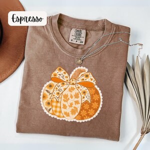 May include: Espresso-coloured t-shirt with a pumpkin graphic. The pumpkin design features a bow and is decorated with sunflowers, leaves, and a gingham pattern. A silver necklace with a pendant is on the shirt. The shirt is labelled Comfort Colors, size S-P-CH.