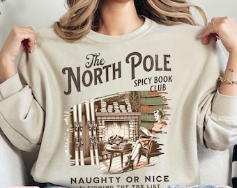 May include: A beige sweatshirt with a graphic design featuring a cabin with a fireplace, a woman reading a book, and the text "The North Pole Spicy Book Club Naughty or Nice Sleigthing the TBR List".