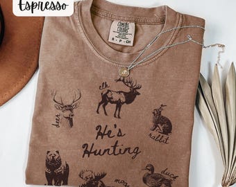 May include: Espresso-colored t-shirt with a crew neck, featuring illustrations of various animals and the text "He's Hunting." A silver necklace with a pendant rests on the shirt. The shirt is labeled "Comfort Colors" and is size small.