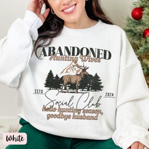 May include: White sweatshirt with a graphic print of a deer in a forest scene. The text reads "Abandoned Hunting Wives Social Club Est. 2024 Hello hunting season, goodbye husband."