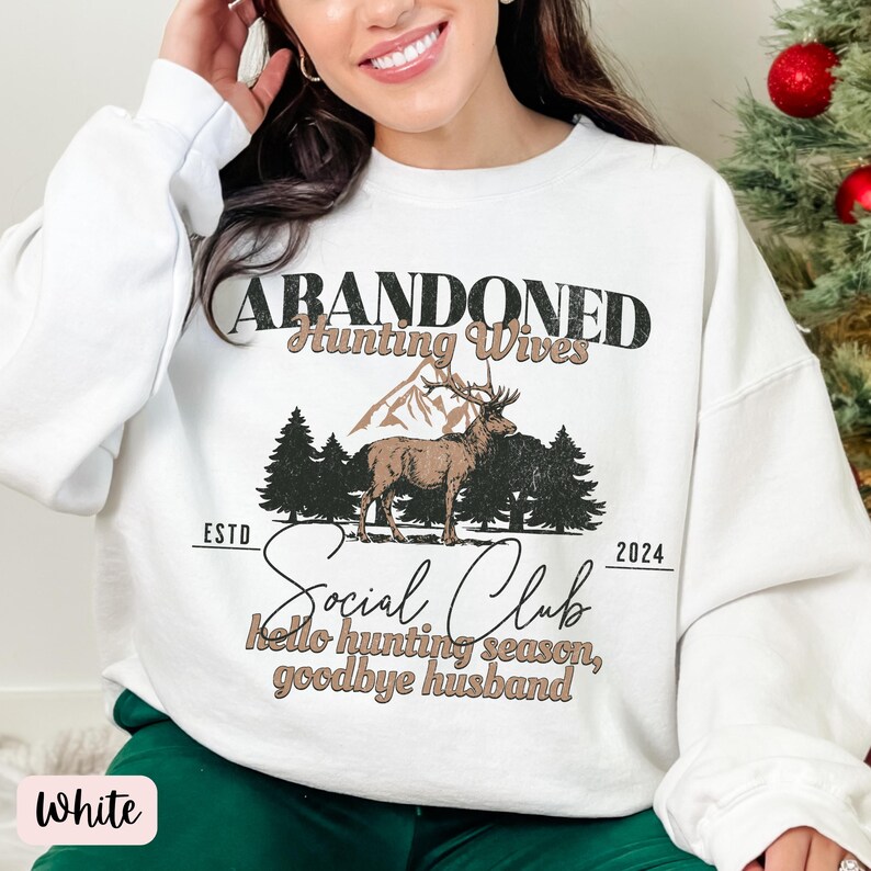 May include: White sweatshirt with a graphic print of a deer in a forest scene. The text reads "Abandoned Hunting Wives Social Club Est. 2024 Hello hunting season, goodbye husband."