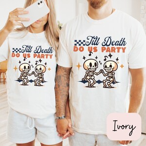 May include: Two white t-shirts with a skeleton couple dancing and the text "Till Death Do Us Party". The shirts are on a couple holding hands. The text is in a retro font with a checkered flag design. The background is a light wood floor.