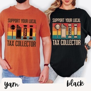 May include: Two t-shirts, one yam-colored and one black, each featuring the text "SUPPORT YOUR LOCAL TAX COLLECTOR." The design includes a lollipop, a chocolate bar, a zig-zag, and a wrapped candy, set against a striped background.