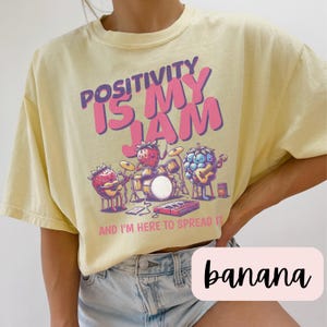 May include: A yellow t-shirt with a graphic of fruit playing instruments. The text on the shirt reads "Positivity is my jam and I'm here to spread it".
