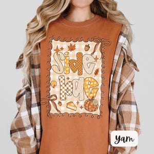 May include: A burnt orange t-shirt with a fall-themed graphic. The graphic features the words "Side Bae" in a mix of fonts and patterns, surrounded by autumn leaves, acorns, and a pumpkin. The shirt is paired with a brown plaid flannel.