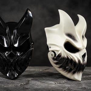 Peut inclure: Deux masques intégraux dotés de protubérances pointues en forme de cornes et de sourires dentés irréguliers. Un masque est fini en noir brillant uni, tandis que l'autre est blanc cassé avec des ombres sombres aérographes autour des yeux et des dents.