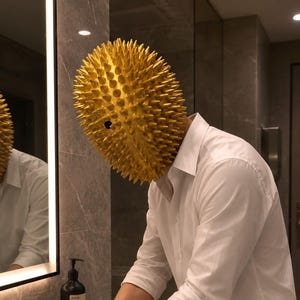 Peut inclure: Un masque ovale doré recouvert de pointes acérées avec une unique ouverture sombre pour l'œil, porté avec une chemise blanche à boutons. Le masque présente une texture dense de pointes saillantes sur toute sa surface, créant une silhouette.