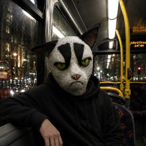 May include: A full-head cat mask featuring white fur texture, black triangular markings above the eyes, and bright green irises. The mask has pointed ears, a pink nose, and a neutral expression. It is worn with a plain black hooded sweatshirt.