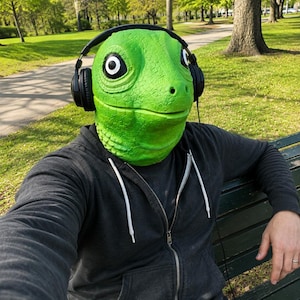 Op de afbeelding: Een felgroen hagedismasker met een getextureerde huid en grote zwart-witte ogen, gedragen met een zwarte hoofdtelefoon. Het masker heeft een realistische reptielvorm met een duidelijke snuit en neusgaten, gecombineerd met een grijze hoodie met rits.