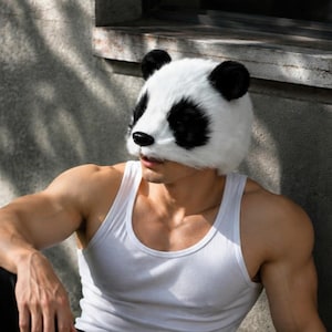 May include: A panda mask in white and black with black ears. The person is wearing a white vest and black trousers. The person is sitting on grass near a concrete wall.