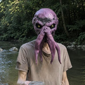 May include: A purple cephalopod-style mask with yellow eyes and dangling tentacles, worn with a plain tan short-sleeved t-shirt. The mask features a textured, wrinkled surface and a prominent forehead, while the t-shirt has a distressed hem.