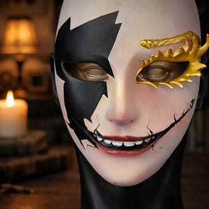 May include: A theatrical mask with a split design. One side is black, the other is pale with a gold eye mask. The mouth is a wide, cracked smile. The mask is on a black stand, with a candle and lamp in the background.
