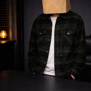 K&ouml;nnte beinhalten: Eine Person, die eine braune Papiert&uuml;te mit zwei Augenl&ouml;chern &uuml;ber dem Kopf, ein gr&uuml;n-schwarz kariertes Hemd und ein wei&szlig;es T-Shirt tr&auml;gt. Die Person steht vor einem schwarzen Tisch.