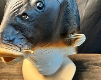 May include: A detailed fish mask with a realistic design. The mask features a dark gray head with textured scales, a prominent eye, and a beige mouth. The mask is mounted on a light beige stand, showcasing the fish's gills and fins.