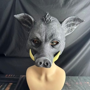 May include: A gray boar mask with yellow tusks and realistic details. The mask has large ears and a textured surface, and is displayed on a mannequin head. The eyes are visible through the eye holes.