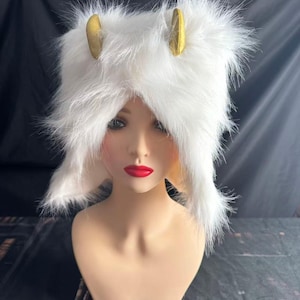 May include: A white faux fur hat with two gold-coloured horns. The hat is fluffy and covers the head and ears. The hat is displayed on a mannequin head with red lipstick.