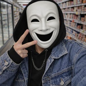 May include: A white smiling mask with black eye holes is worn over a black hooded sweatshirt and a denim jacket. A silver chain necklace is visible. The person is giving a peace sign.