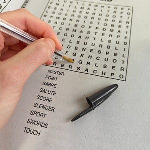 May include: A crossword puzzle titled "ON GUARD" with a clear pen and a black pen cap. The puzzle is partially filled in, with a list of words below. The pen is held by a person.