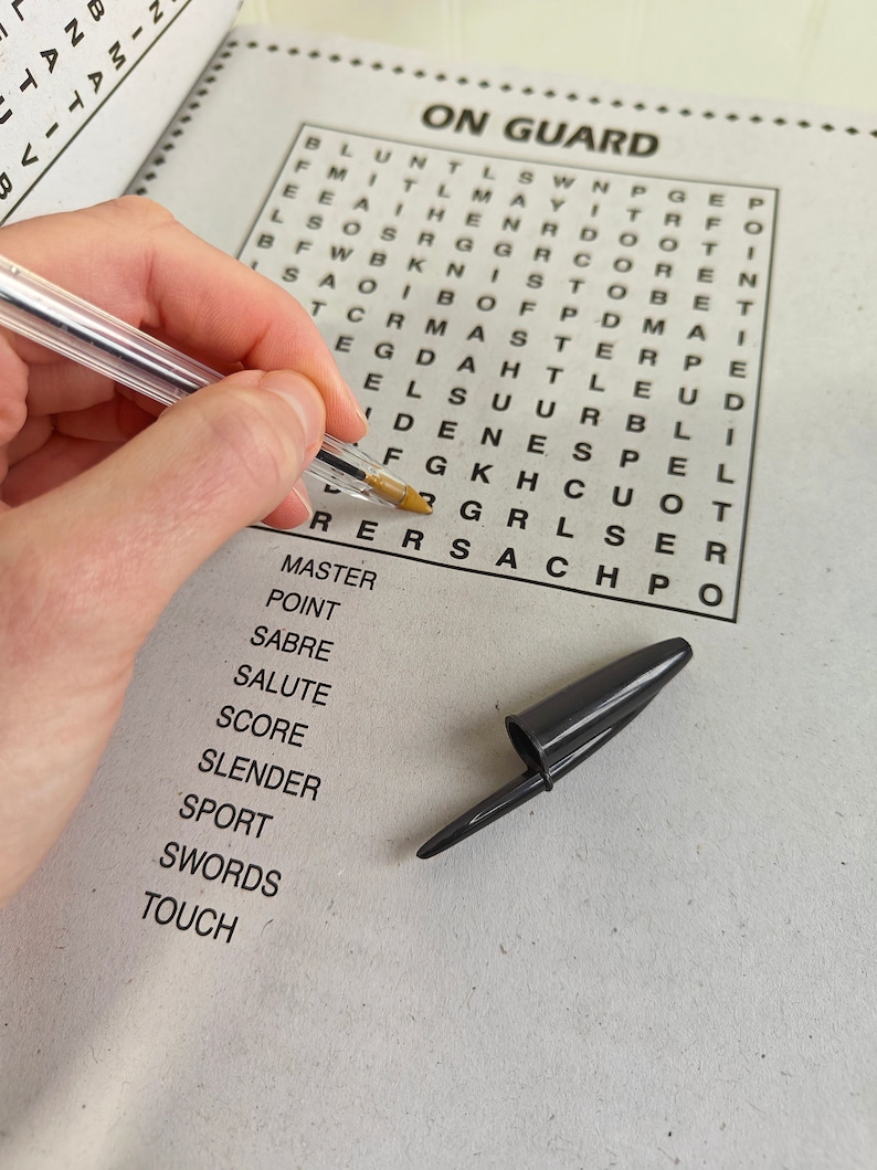 May include: A crossword puzzle titled "ON GUARD" with a clear pen and a black pen cap. The puzzle is partially filled in, with a list of words below. The pen is held by a person.