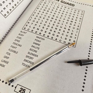 May include: Open word search puzzle book with the title "ON GUARD" and a clear pen resting on the page. The puzzle is in black text on a white background. The number 25 is in a box at the bottom of the page.