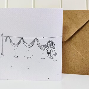 A square greetings card with a brown recycled envelope behind standing on a white table. A black fine line illustration of an older man relaxing in a chair, reading a book whilst drying his long moustache on a straight clothes line outside.