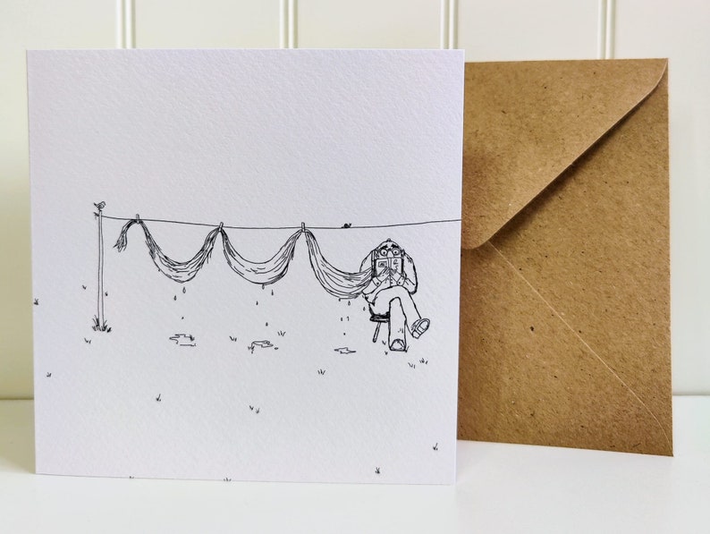 A square greetings card with a brown recycled envelope behind standing on a white table. A black fine line illustration of an older man relaxing in a chair, reading a book whilst drying his long moustache on a straight clothes line outside.