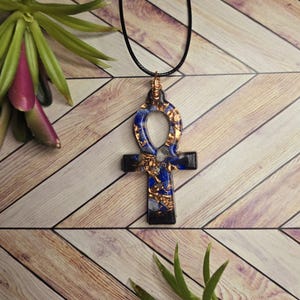 May include: An ankh pendant necklace featuring a black resin base embedded with blue stone fragments and metallic copper flakes. The pendant is suspended from a thin black cord and secured at the top with a wrapped copper wire bail.