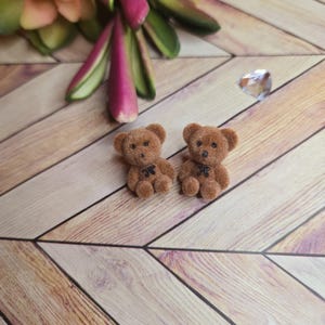 May include: Two small brown teddy bears with black bow ties sit on a wooden surface. The bears have small black eyes and are made of a soft, fuzzy material. A small, clear crystal and a colorful floral arrangement are in the background.