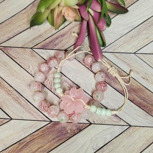 Puede incluir: Dos pulseras de cuentas rosadas y verdes con un dije de flor rosa. Las pulseras están sobre una superficie de madera.