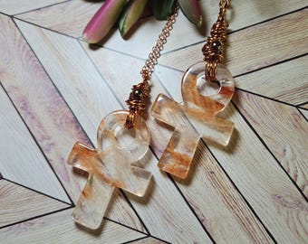 May include: A pair of ankh-shaped earrings. The earrings are made of clear resin with orange and white veining. They hang from copper-colored chains and are accented with copper wire and beads. The background features a wooden surface and greenery.