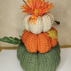 Op de afbeelding: Een stapel van drie gehaakte pompoenen, de bovenste is versierd met een grote oranje bloem. De pompoenen zijn beige, oranje en groen.