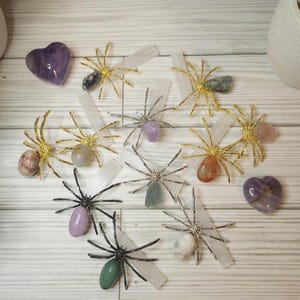 May include: A collection of eight gold and silver spider sculptures with various colored gemstones in the center of each spider. The spiders are arranged on a white wooden surface with selenite wands.