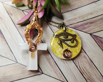 May include: Two gold necklaces with pendants. One pendant is a white resin Ankh with gold accents. The other pendant is a round resin piece with a gold Eye of Horus design.