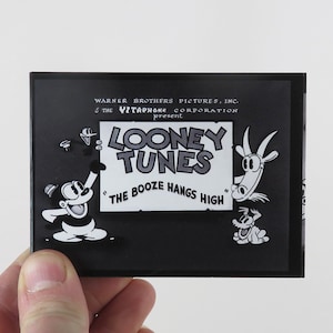 Może przedstawiać: Czarno-biała ilustracja postaci z kreskówki trzymającej znak z napisem "Looney Tunes" i "The Booze Hangs High". Znak jest otoczony innymi postaciami z kreskówek.