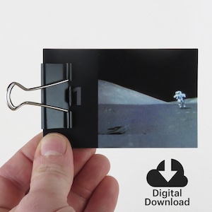 Könnte beinhalten: Ein Schwarzweißfoto eines Astronauten, der auf dem Mond geht. Das Foto ist auf einer Karte mit schwarzem Rand gedruckt, auf der links die Zahl "1" steht. Die Karte wird von einer Hand gehalten, an der oben ein Büroklammer befestigt ist.