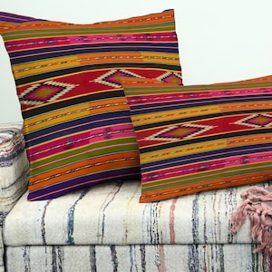May include: Two decorative throw pillows with a vibrant, striped pattern in red, orange, yellow, pink, and purple. The pillows feature diamond-shaped accents and are placed on a patterned sofa, creating a colorful and inviting aesthetic.