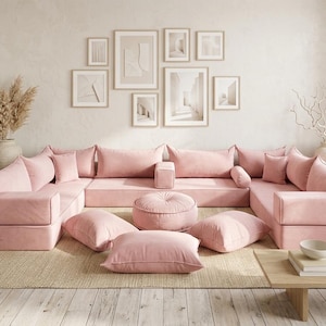 May include: A modular pink sectional sofa with multiple cushions and a round ottoman. The sofa is arranged on a neutral-colored rug, with framed artwork on the wall behind it. Decorative vases and a small wooden table complete the living room setting.