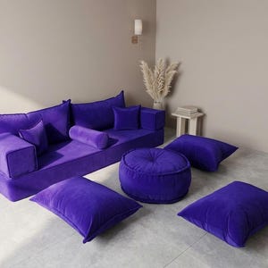 May include: A modern, modular sofa set in a vibrant purple hue. The set includes a large sofa, a round ottoman, and several square and cylindrical pillows. The plush velvet fabric adds a touch of luxury to this contemporary living room furniture.