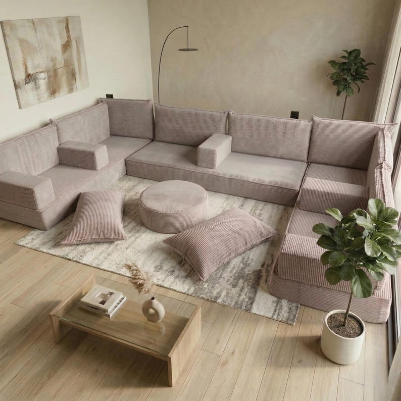 May include: A modular sectional sofa in a light pink corduroy fabric. The set includes multiple seat cushions, armrests, and throw pillows. A round ottoman and a rectangular coffee table with a glass top are also present. The room is decorated with a rug and plants.
