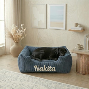 以下が含まれることがあります： 前面に「Nakita」と刺繍された青いコーデュロイの犬用ベッド。黒い犬がベッドの中で休んでいます。ベッドはニュートラルなトーンの装飾が施された部屋にあります。